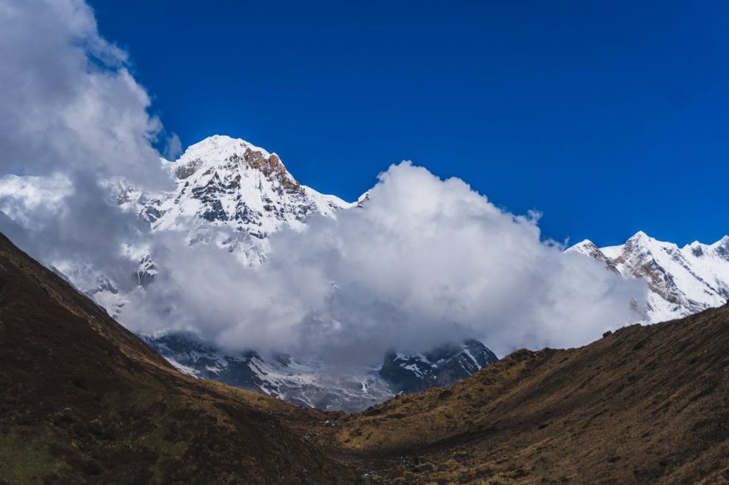 a-mountain-range-with-clouds-and-blue-sky-26570336 Spectacular view of snow-capped Machhapuchchhre peak surrounded by clouds in Nepal.